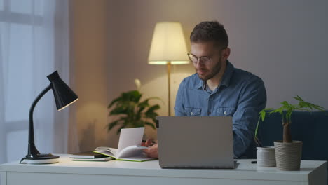 Männlicher-Student-Lernt-Abends-Zu-Hause-Und-Liest-Notizen-Im-Notizbuch-Und-Informationen-Im-Internet-Mit-Dem-Laptop,-E-Learning-Und-Berufliche-Weiterbildung